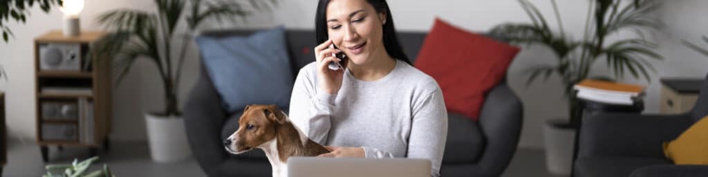 Woman talking on the phone looking at a computer petting a white dog with brown spots